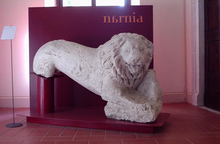 Stone sculpture of a crouching lion, displayed on a red pedestal in front of the inscription Narnia at Palazzo Eroli.