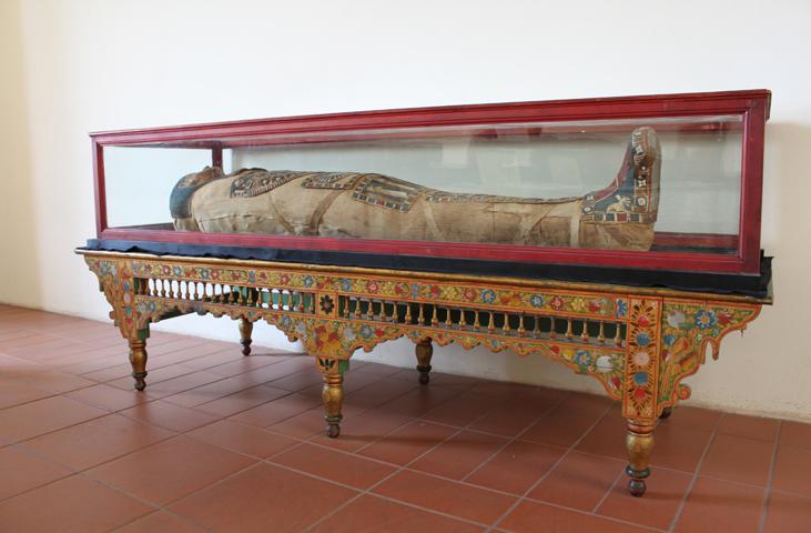 Bandaged mummy displayed in a glass case on a decorated wooden table at Palazzo Eroli.